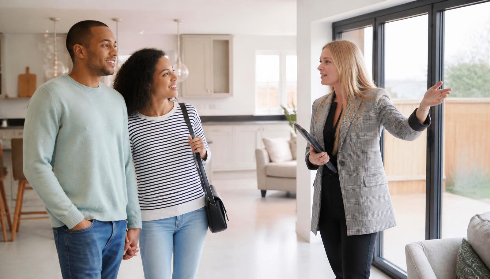 A young couple touring a bright, modern home with a real estate agent pointing toward the windows